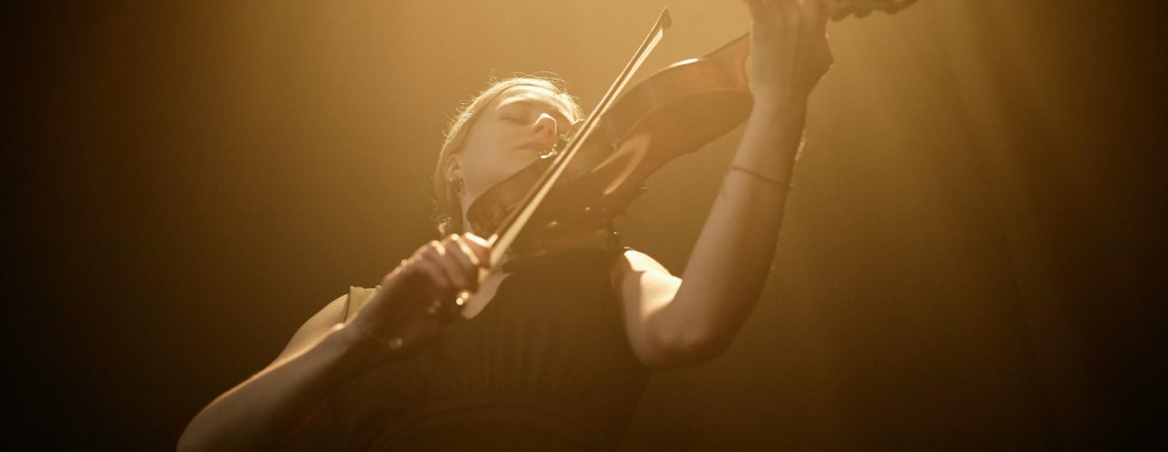 Low angle shot of female classical music artist sensually playing violin illuminated by light from above in dark studio, copy space